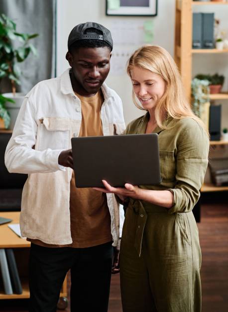Two young intercultural employees in casualwear looking at screen of laptop held by blond businesswoman during discussion
