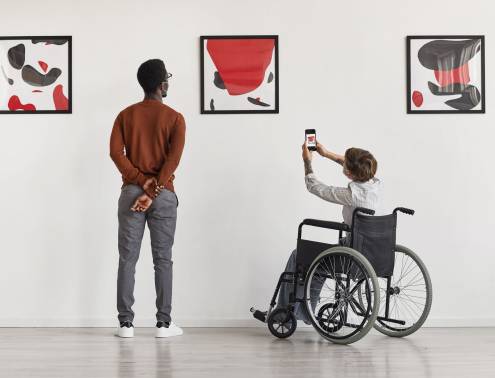 Back view portrait of young woman using wheelchair taking photo of artwork while visiting accessible museum, copy space
