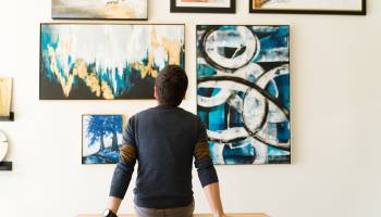 Hispanic male visitor looking reflective while sitting on a bench and admiring the various paintings on the wall of an art gallery