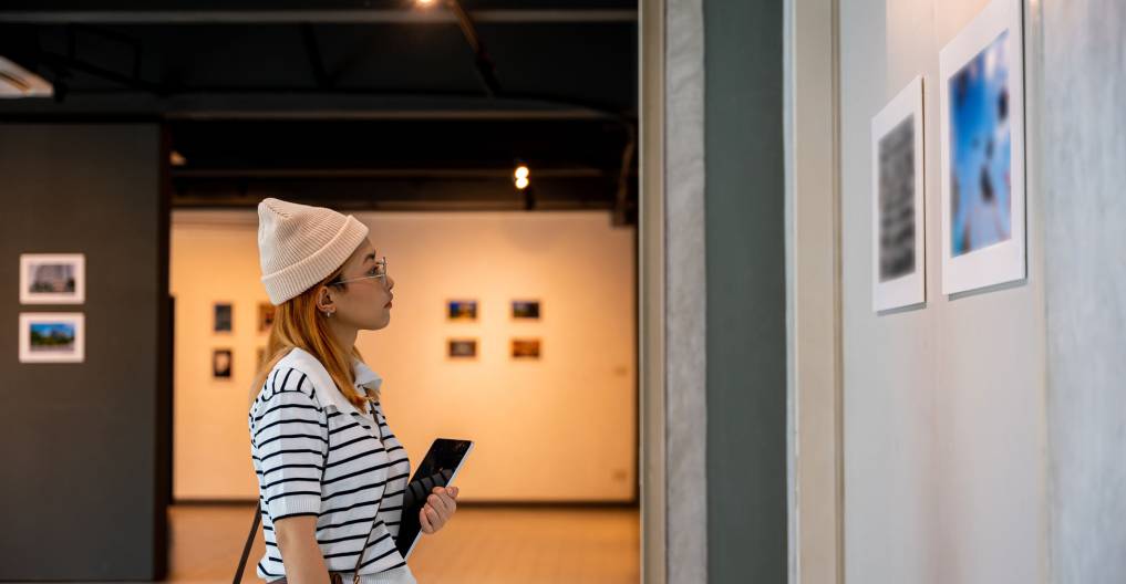 Young person at photo frame hold digital book leaning against at show exhibit artwork gallery, Asian woman holding tablet at art gallery collection in front framed paintings looking pictures on wall